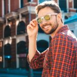 Handsome smiling stylish hipster lambersexual model.Modern man dressed in red checkered shirt. Fashion male posing near skyscraper on the street background in sunglasses. Outdoors at sunset - Hidalgo