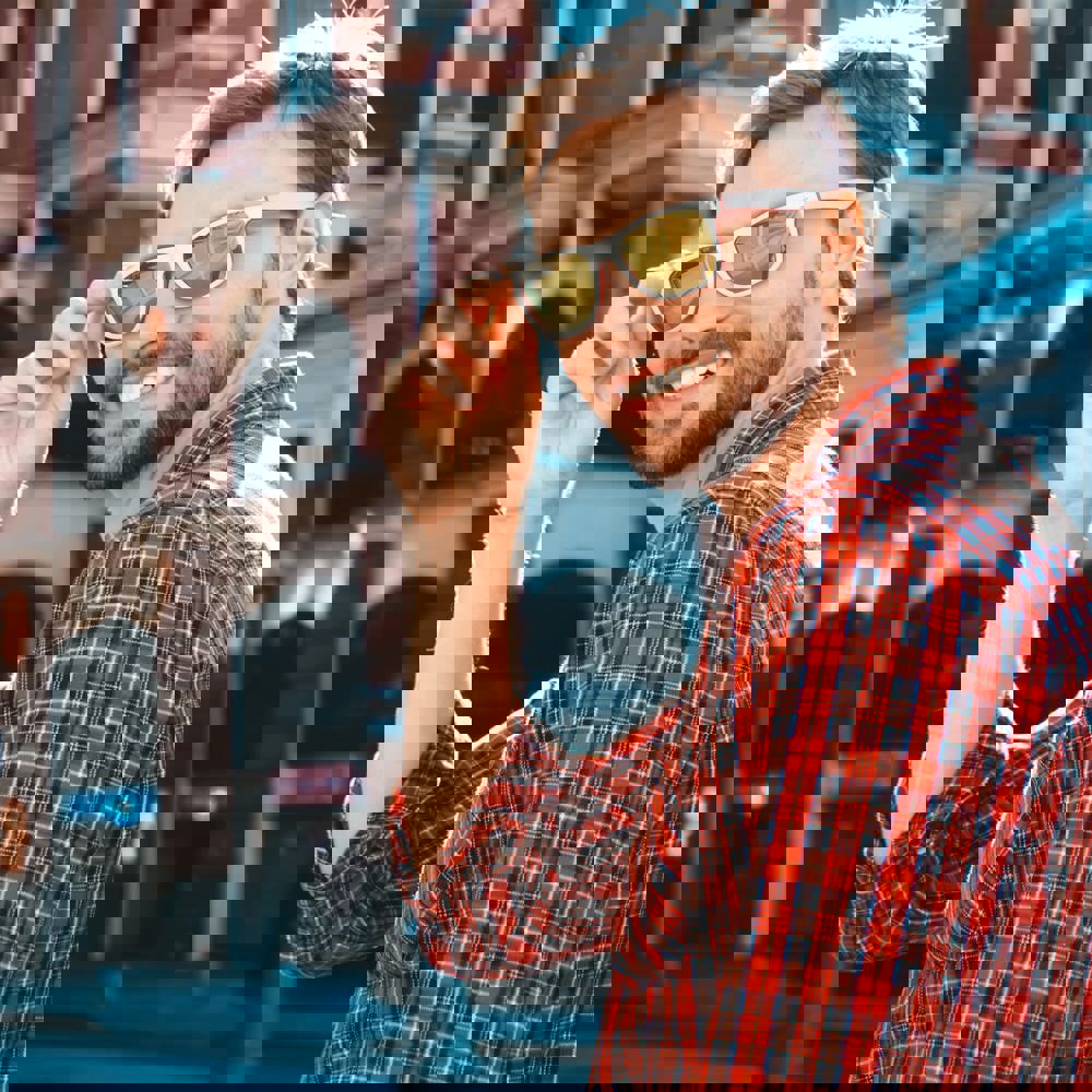 Handsome smiling stylish hipster lambersexual model.Modern man dressed in red checkered shirt. Fashion male posing near skyscraper on the street background in sunglasses. Outdoors at sunset - Hidalgo