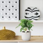 White shelf display with a geometric black-and-white wall print on the left, a round black-and-white striped plate propped on the right, a green monstera plant in a ribbed white pot in the center, and a gold wavy decorative vase on a wooden surface to the left.