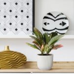 Decorative interior vignette: potted plant on a wooden surface with a gold wavy vase and a round plate with a black abstract face beside geometric black-and-white wall art.