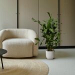 Beige textured armchair beside a tall leafy green plant in a white pot on a round jute rug, against a neutral cabinet backdrop in a modern living room.