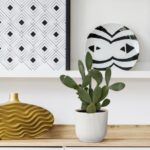 Decorative still life with a ribbed white planter cactus on a wooden surface, a gold wavy vase, and black-and-white geometric art plate on a white shelf.
