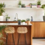 Modern kitchen with a wooden island, two wicker bar stools, and numerous potted plants on the counter and shelves.