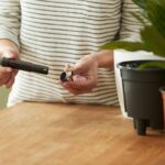 Person in a striped shirt attaches a small cap to a white and black kitchen appliance on a wooden table.