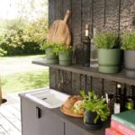 Outdoor patio kitchen shelf with potted herbs, a cutting board, bottles, and a cookbook on a dark wooden counter. In the background, a sunny garden.