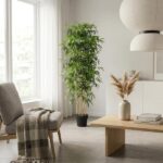 Bright living room corner with a tall potted plant, gray chair with a blanket, and a wooden coffee table under a large white pendant lamp.