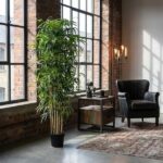 Industrial-style lounge with a tall potted plant by large grid windows, a black armchair, a small table holding a mug and vintage camera, and a patterned rug.