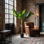 Industrial loft seating area with exposed brick walls, a tall potted banana plant, and a brown leather armchair by large black-framed windows.