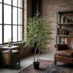Industrial-style living room with a tall potted tree next to a large window and a brown leather armchair nearby by a metal bookshelf.