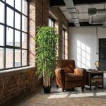 Industrial loft corner with large window, tall potted plant, and brown leather armchair beside a wooden side table.