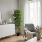 Bright living room with a tall potted plant, white sideboard, gray armchair, and a beige throw near sheer-curtained windows.