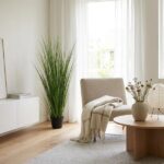 Bright, minimalist living room with a tall potted plant, beige chair, and a round wooden coffee table by a window with sheer white curtains.