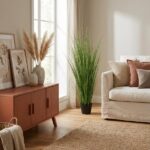 Living room corner with a mid-century wooden cabinet, framed botanical prints, pampas grass vases, and a beige sofa with cushions by a window.
