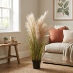 Tall pampas grass in a black pot sits on a woven rug beside a beige sofa with rust and cream cushions in a sunlit living room.