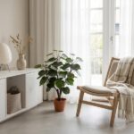Bright, airy living room with a potted rubber plant, light wood chair with a blanket, and sheer white curtains by a large window.