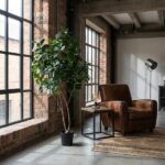 Industrial loft living room: brown leather armchair, small side table, tall potted plant, and large brick windows.