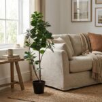Tall potted plant beside a beige sofa in a bright living room with a wooden side table, mug, and woven rug by the window.