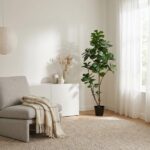 Cozy living room corner with a gray armchair, beige throw, white cabinet, and a tall leafy plant by a sheer-curtained window.