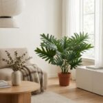 Cozy living room with a large potted tropical plant by a window, beige sofa, and a round wooden coffee table with dried stems in a vase.