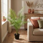 Bright living room with a tall potted palm beside a window, a beige sofa with orange and green cushions, and a wooden sideboard in the background.
