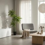 Bright living room with a gray lounge chair, beige throw, tall potted plant, white cabinet, and a large window letting in light from behind sheer curtains; a wooden coffee table sits nearby.