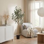 Tall potted indoor tree beside a light gray armchair in a cozy beige living room with a white cabinet, blanket, and natural light streaming through a window.