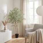 Bright living room with a tall potted tree, beige curtains, and a gray armchair with a knit throw by a window.
