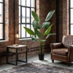 Industrial living room with exposed brick walls, large windows, a tall green potted plant, a wooden-and-metal side table with a glowing desk lamp, a brown leather armchair, and a patterned rug.