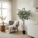 Cozy living room corner with a gray upholstered armchair, cream blanket, round wooden side table with a vase of pampas grass, and a tall potted leafy plant by a white cabinet.