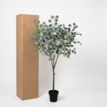 Artificial eucalyptus plant in a black pot beside a tall cardboard box on a white background.