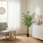 Bright living room corner with a gray armchair, beige throw, white sheer curtains, and a tall potted palm beside a white cabinet.