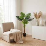 Beige armchair with a cream knit blanket beside a tall potted monstera plant in a bright, minimalist living corner.