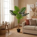 Tall potted indoor plant with large green leaves beside a beige sofa in a sunlit living room. A wicker basket and small vases sit on a wooden bench nearby.