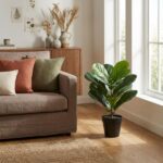 Cozy living room corner with beige sofa, rust and green cushions, wooden sideboard, and a large potted plant by a sunlit window.