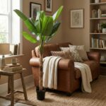 Tall potted green plant beside a brown leather sofa in a sunlit living room with knit throws and cushions on a tan rug.