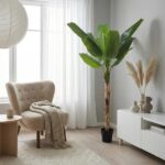 Cozy living room nook with beige armchair, knit throw, small side table, tall potted plant, white curtains, and a white cabinet with vases.