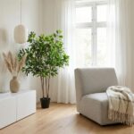 Bright minimalist living room with a large potted plant, white curtains, a gray armchair with a beige throw, and a white cabinet with a vase of dried grasses.