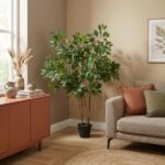 Tall potted tree in a living room corner beside a peach cabinet and beige sofa with orange and green cushions; framed fern print on the wall.