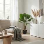 Bright living room with a beige sofa, plaid throw, and a light wooden coffee table; a large potted plant stands by a white cabinet with a vase and dried grasses; sun streams through sheer curtains.