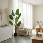 Bright minimalist living room with a beige armchair, cream throw, tall leafy plant, and light wood coffee tables.