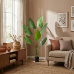 Sunlit living room with a tall potted tropical plant beside a beige sofa, framed botanical art, and a wooden console with clay pots.
