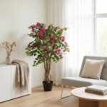 Bright living-room corner with a tall potted pink flowering plant beside a gray chair, round wooden coffee table, beige pillow, and sheer-curtained window.