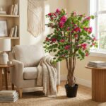 Cozy beige living room corner with a chair, knitted throw, woven wall art, and a large potted flowering plant with pink blossoms beside a round side table.