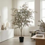 Tall indoor potted tree in a bright minimalist living room with white furniture and sheer curtains.