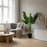 Cozy living room corner with a gray armchair, beige knit throw, round wood coffee table, and a tall potted plant by the window.