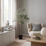 Tall potted olive tree in a light-filled living room with a beige armchair, grey cushion, and natural wood coffee table.