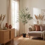 A cozy living room with a tall potted olive tree beside a window, a beige sofa with rust and cream cushions, and a wooden sideboard with ceramic vases.