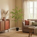 A cozy living room with a potted tree, beige walls, and a neutral sofa with orange, cream, and green cushions by a sunlit window.