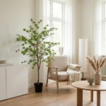 Bright minimalist living room corner with a tall potted plant, beige armchair, round wooden coffee table, and floor lamp in natural light.