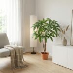 Tall leafy houseplant in a terracotta pot beside a white floor lamp in a minimalist living room with a beige sofa and throw blanket on the left.